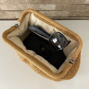 May include: A woven straw bag with a light brown color and a white lining. The bag is open and has sunglasses and a phone inside.
