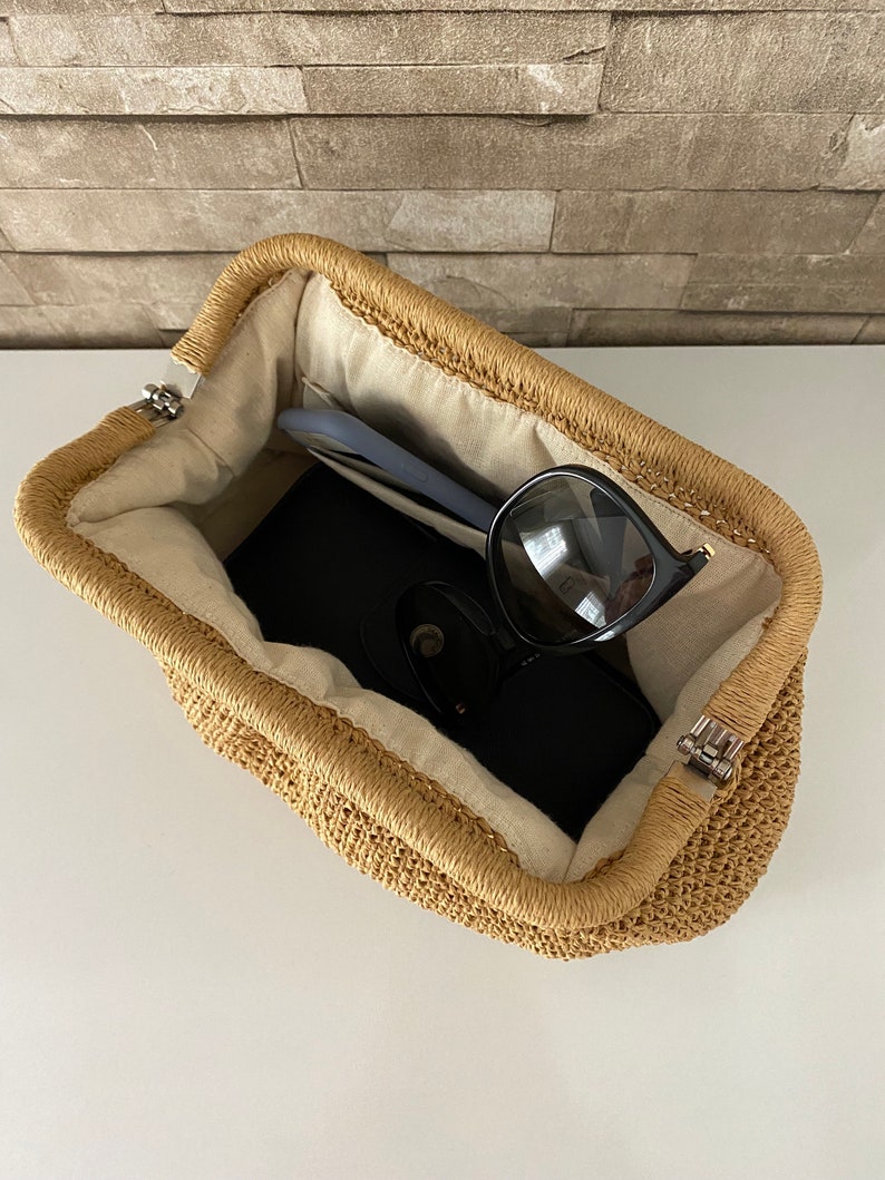 May include: A woven straw bag with a light brown color and a white lining. The bag is open and has sunglasses and a phone inside.