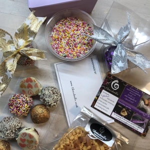 May include: A gift box filled with chocolate truffles, sprinkles, and other treats. The box is decorated with a silver and gold ribbon. The box includes a card that says "Handmade by Chocadoodle" and a website address.
