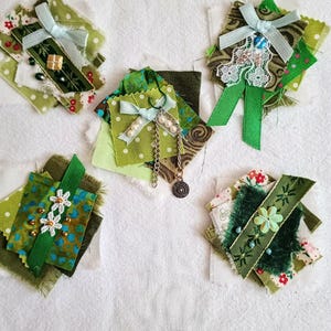 May include: Five fabric embellishments in shades of green, featuring layered textile scraps, light blue ribbons, white lace, small beads, metallic chains, and floral patterns. Each piece is constructed from various fabric textures and decorative trims.