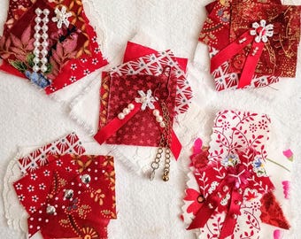May include: Five fabric patches in shades of red and white, featuring layered textile scraps, floral and geometric patterns, white lace accents, pearl beads, red ribbons, metallic studs, and a small brass-toned chain with a bell charm.