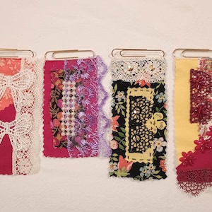 Puede incluir: Cuatro marcapáginas de tela hechos a mano con clips. Cada marcapáginas presenta un diseño único con encaje, estampados florales y elementos decorativos en tonos de rojo, rosa, amarillo y negro. Los marcapáginas son rectangulares y tienen una variedad de texturas.