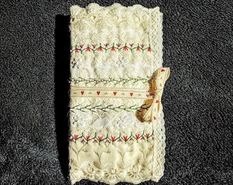 May include: A rectangular cream-colored fabric cover featuring intricate white lace borders, embroidered floral patterns in red and green, and a central ribbon tie decorated with a repeating pattern of small red hearts.