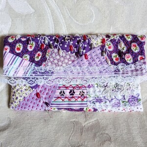 Könnte beinhalten: Eine handgefertigte Patchwork-Clutch in Lila-, Lavendel- und Weißtönen. Der Stoff weist Blumen- und geometrische Muster auf, die mit roten und weißen Perlen und zarten weißen Spitzen verziert sind. Die Clutch ist rechteckig und gefaltet.