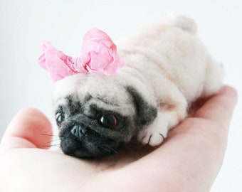 May include: A small, light brown pug puppy wearing a pink bow. The puppy is lying on a person's hand.