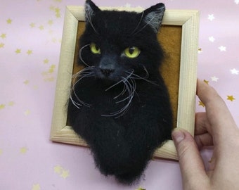 Pode incluir: Um retrato de um gato preto com bigodes brancos feito com feltro de agulha emoldurado. O gato tem olhos amarelos e olha diretamente para o espectador.