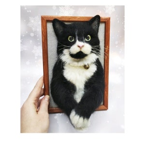 May include: A framed, felted cat portrait. The cat is black and white with a distinguished mustache and a small bell collar. The frame is a warm brown, and the background is a soft gray.