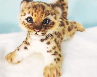 May include: A small, realistic plush toy leopard cub with brown and white fur, blue eyes, and a white belly. The cub is lying on a white surface.