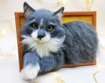 Pode incluir: Uma escultura de gato de feltro cinza e branco em uma moldura de madeira. O gato está deitado com a cabeça virada para o lado e as patas visíveis. O gato tem olhos verdes e um bigode branco.