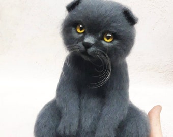 May include: A gray, felted Scottish Fold cat with yellow eyes, sitting on a hand. The cat has a long tail and is looking to the left.