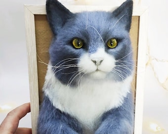 Pode incluir: Um gato cinza e branco com olhos amarelos é emoldurado em uma moldura de madeira clara. O gato é feito de feltro e tem uma aparência 3D.