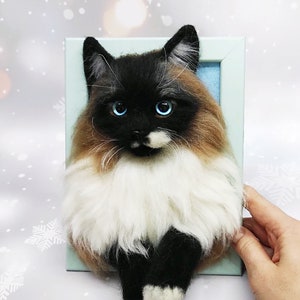 Könnte beinhalten: Ein Nadelfilz-Katzenporträt in einem hellblauen Rahmen. Die Katze ist ein Siam mit braunem, schwarzem und weißem Fell. Die Katze hat blaue Augen und sitzt mit den Pfoten unter dem Körper.