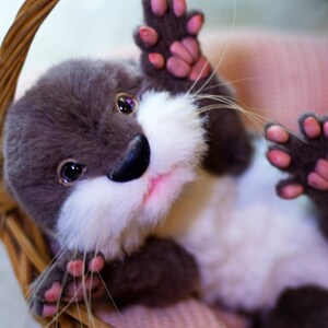 Op de afbeelding: Een grijze en witte pluche otter met roze pootjes zit in een rieten mand. De otter heeft een witte snuit en een roze neus.