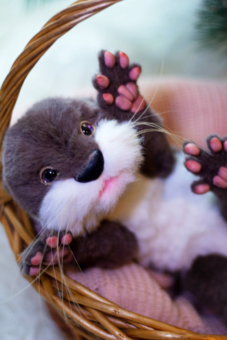 Op de afbeelding: Een grijze en witte pluche otter met roze pootjes zit in een rieten mand. De otter heeft een witte snuit en een roze neus.