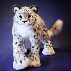 May include: A white and gray plush snow leopard with black spots. The snow leopard is standing on a blue surface and has its head turned to the left.
