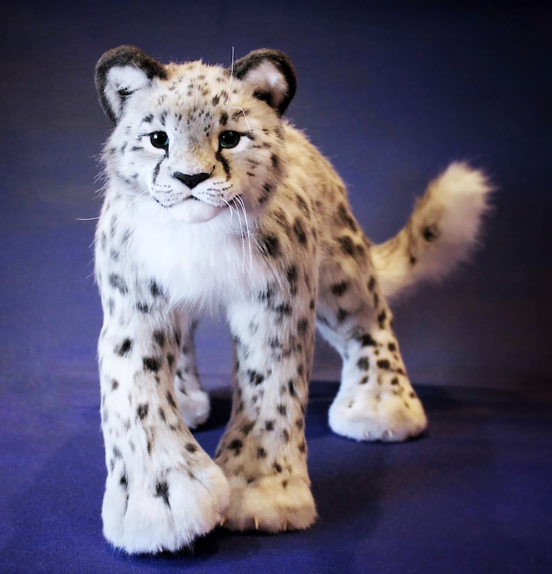 May include: A white and gray plush snow leopard with black spots. The snow leopard is standing on a blue surface and has its head turned to the left.