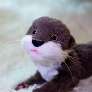 Op de afbeelding: Een pluche otter met bruine vacht, witte vacht op zijn borst en een roze geverfde neus en mond. De otter ligt op een wit, pluche oppervlak.