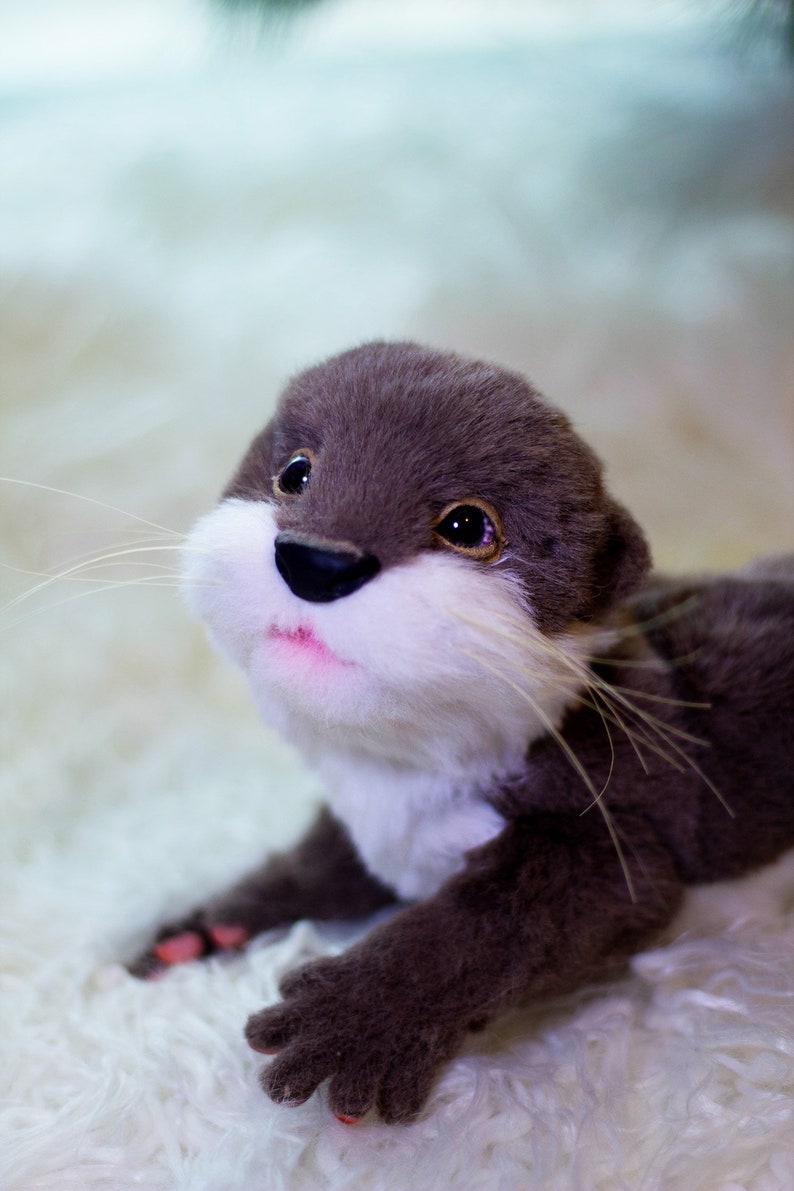 Op de afbeelding: Een pluche otter met bruine vacht, witte vacht op zijn borst en een roze geverfde neus en mond. De otter ligt op een wit, pluche oppervlak.