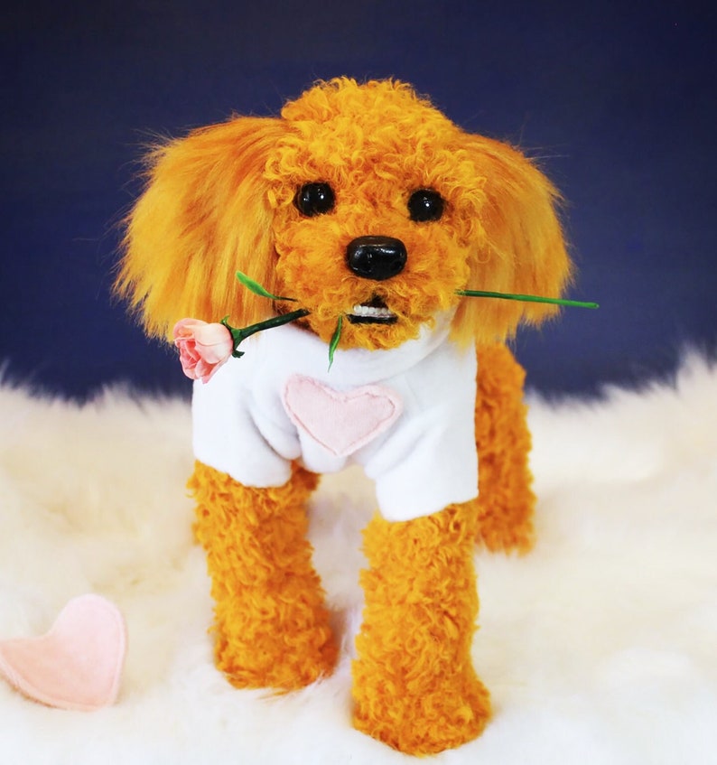 May include: A plush toy poodle with orange fur wearing a white shirt with a pink heart on the front. The poodle is holding a green stem with a pink flower in its mouth.