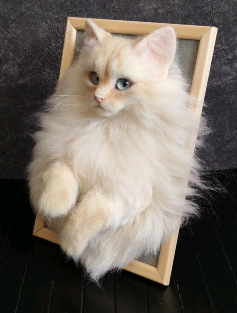K&ouml;nnte beinhalten: Skulptur einer cremefarbenen Katze mit blauen Augen, die aus einer Holzrahmenleinwand zu kommen scheint. Das Fell der Katze ist lang und strukturiert, mit einem realistischen Aussehen. Das Kunstwerk ist vor einem dunklen Hintergrund platziert.
