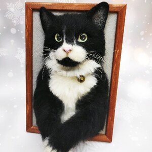 May include: A framed, handcrafted cat portrait. The cat is black and white with green eyes, a pink nose, and a small gold bell on its collar. The portrait is set in a brown wooden frame against a gray background.