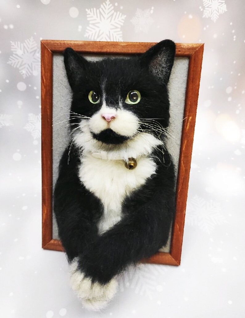 May include: A framed, handcrafted cat portrait. The cat is black and white with green eyes, a pink nose, and a small gold bell on its collar. The portrait is set in a brown wooden frame against a gray background.