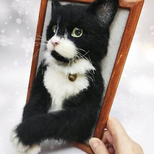 May include: A handcrafted, framed felt cat portrait. The black and white cat has green eyes, a pink nose, and a small gold bell on its collar. The wooden frame is a warm brown color, and the cat is set against a light gray background.