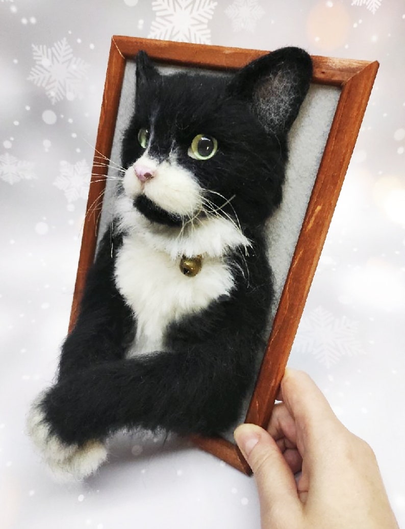 May include: A handcrafted, framed felt cat portrait. The black and white cat has green eyes, a pink nose, and a small gold bell on its collar. The wooden frame is a warm brown color, and the cat is set against a light gray background.