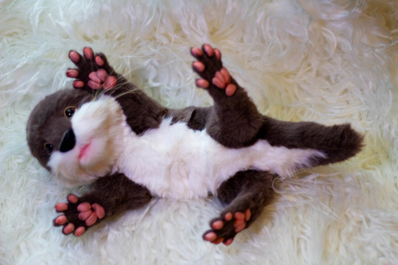 Op de afbeelding: Een pluche otter met bruine vacht, een witte buik en roze pootjes. De otter ligt op zijn rug met zijn poten in de lucht.