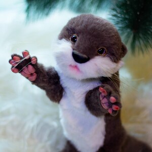 Op de afbeelding: Een pluche otter speelgoed met bruine vacht, een witte borst en roze pootjes. De otter staat op een wit, pluche oppervlak en heeft een speelse uitdrukking.