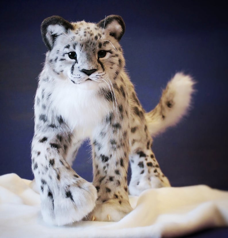 May include: A white and gray plush snow leopard with black spots. The snow leopard is standing on a white surface and has its paws outstretched.