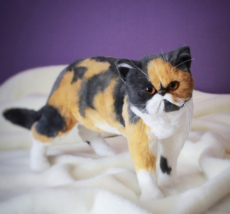 May include: A calico cat with black, orange, and white fur. The cat has a white face with black markings around the eyes and nose. The cat is standing on a white blanket.
