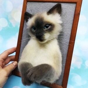 K&ouml;nnte beinhalten: Skulptur einer Siamkatze mit cremefarbenem K&ouml;rper, dunkelbraunem Gesicht, Ohren und Pfoten sowie leuchtend blauen Augen. Die Katze ist auf einem grauen Filzhintergrund in einem Holzrahmen montiert, wodurch ein 3D-Effekt entsteht. Einzigartige Heimdekoration.