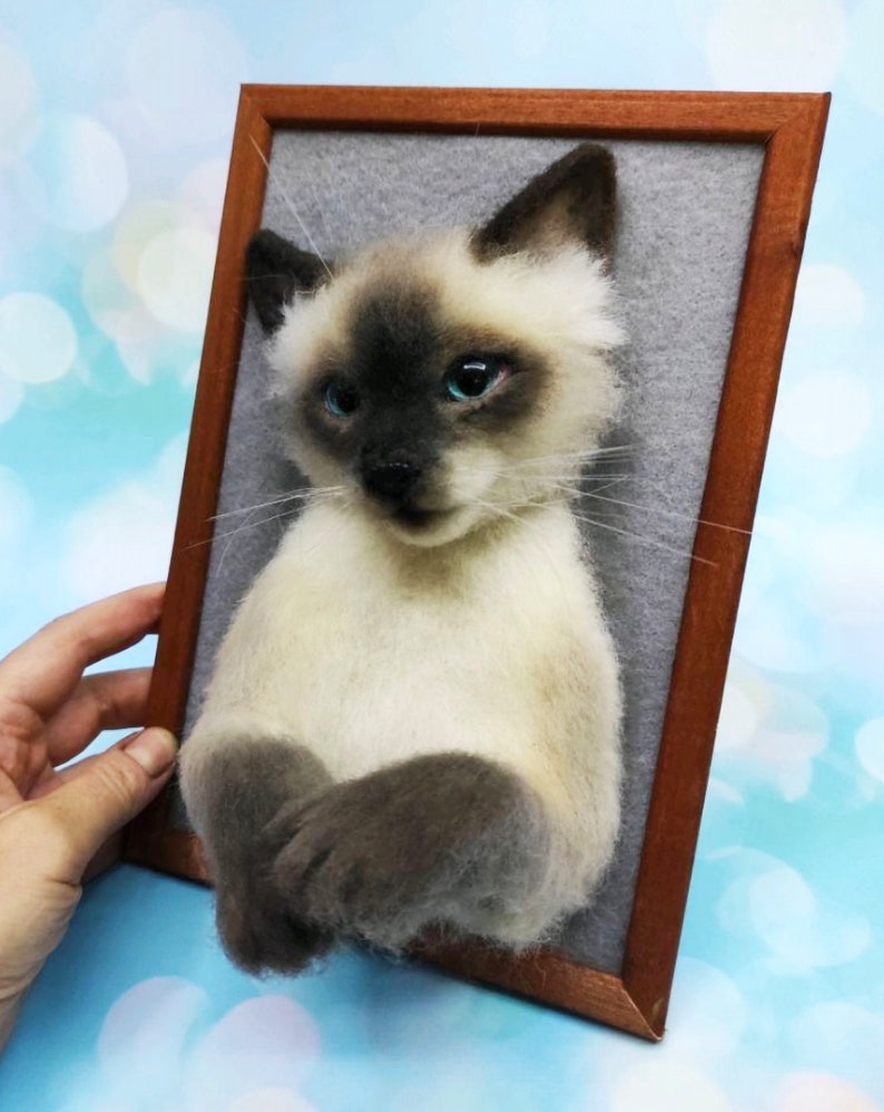 K&ouml;nnte beinhalten: Skulptur einer Siamkatze mit cremefarbenem K&ouml;rper, dunkelbraunem Gesicht, Ohren und Pfoten sowie leuchtend blauen Augen. Die Katze ist auf einem grauen Filzhintergrund in einem Holzrahmen montiert, wodurch ein 3D-Effekt entsteht. Einzigartige Heimdekoration.
