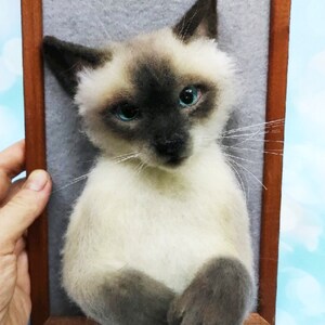K&ouml;nnte beinhalten: Gefilzte Skulptur einer Siamkatze in einem Holzrahmen. Die Katze hat einen cremefarbenen K&ouml;rper, dunkelbraune Ohren und ein Gesicht sowie auff&auml;llige blaue Augen. Der Rahmen hat eine warme braune Farbe, der Hintergrund ist hellgrau.