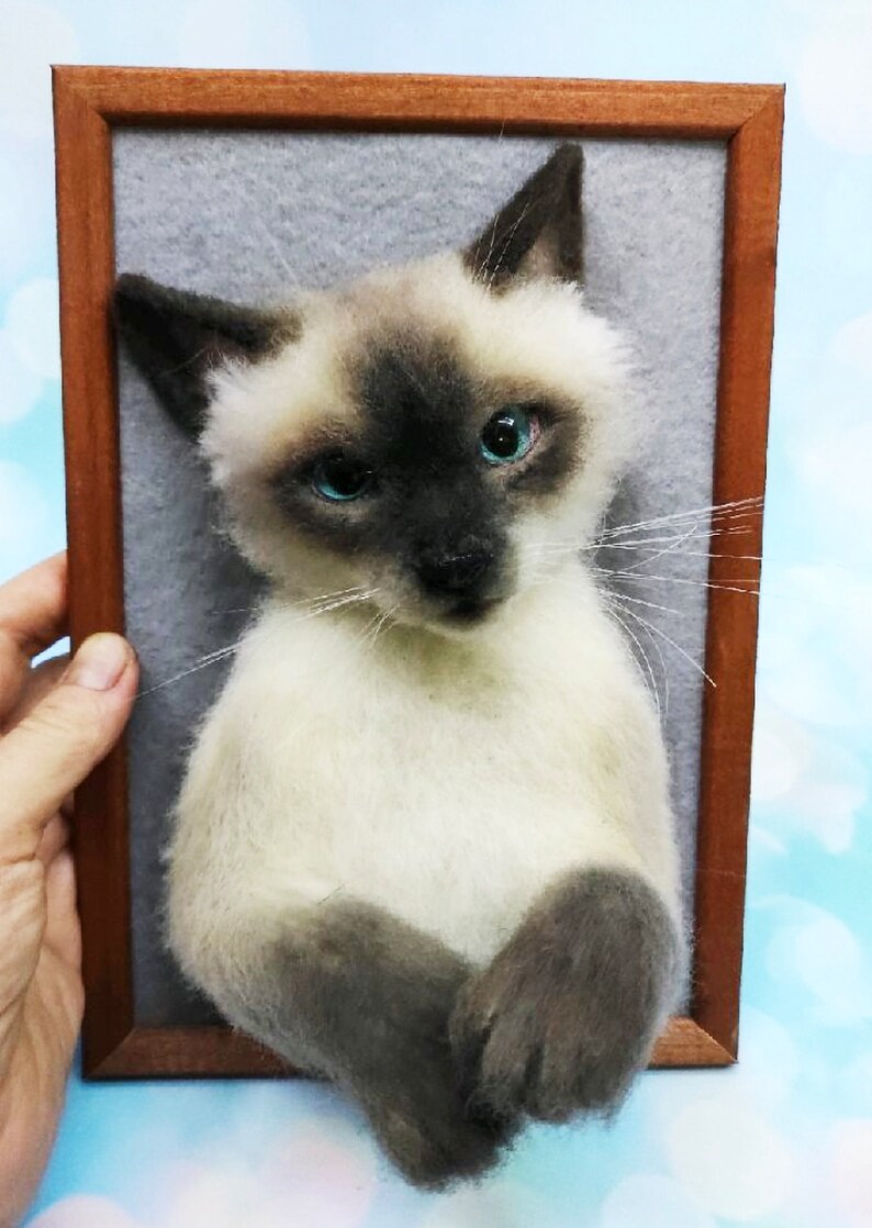 K&ouml;nnte beinhalten: Gefilzte Skulptur einer Siamkatze in einem Holzrahmen. Die Katze hat einen cremefarbenen K&ouml;rper, dunkelbraune Ohren und ein Gesicht sowie auff&auml;llige blaue Augen. Der Rahmen hat eine warme braune Farbe, der Hintergrund ist hellgrau.