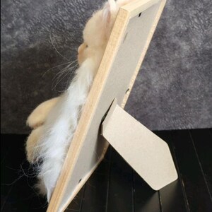 K&ouml;nnte beinhalten: Handgefertigte, hellbeige gefilzte Katzenfigur mit langen wei&szlig;en Schnurrhaaren und einem langen wei&szlig;en Bart, pr&auml;sentiert in einem hellen Holzbilderrahmen mit einem Karton-Staffelei-R&uuml;cken. Die Katze befindet sich links vom Rahmen.