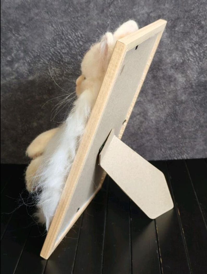 K&ouml;nnte beinhalten: Handgefertigte, hellbeige gefilzte Katzenfigur mit langen wei&szlig;en Schnurrhaaren und einem langen wei&szlig;en Bart, pr&auml;sentiert in einem hellen Holzbilderrahmen mit einem Karton-Staffelei-R&uuml;cken. Die Katze befindet sich links vom Rahmen.