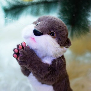 Op de afbeelding: Een pluche bruine en witte otter met een witte borst en een roze neus. De otter houdt zijn poten bij elkaar in een gebedshouding.