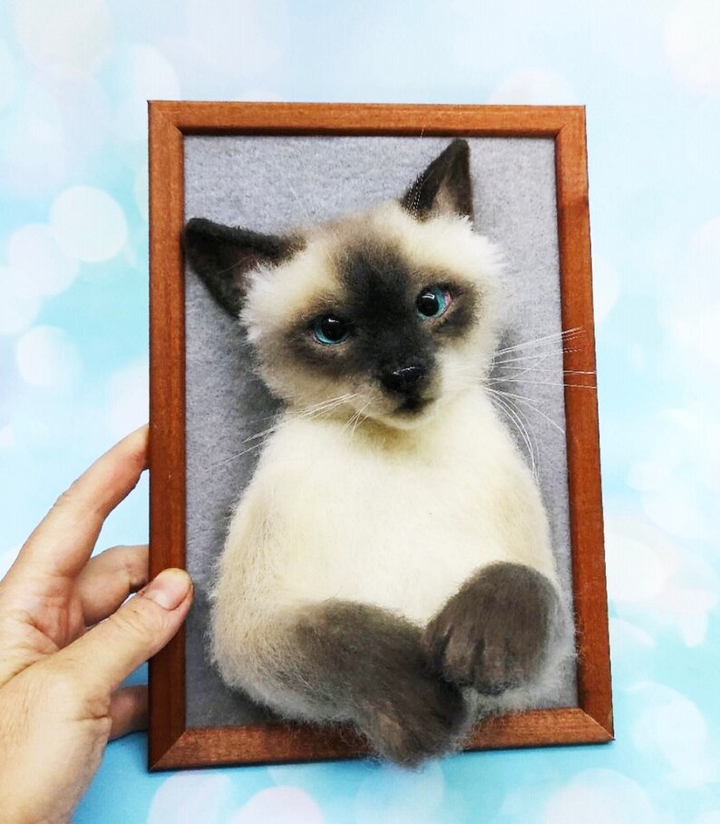 K&ouml;nnte beinhalten: Ein gerahmtes Portr&auml;t einer Siamkatze. Die Katze hat einen cremefarbenen K&ouml;rper, dunkelbraune Punkte und leuchtend blaue Augen. Der Holzrahmen ist warmbraun, vor einem weichen, grauen Hintergrund.