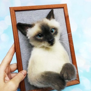 K&ouml;nnte beinhalten: Ein gerahmtes Portr&auml;t einer Siamkatze. Die Katze hat cremefarbenes Fell mit dunkelbraunen Punkten und blauen Augen. Der Holzrahmen hat eine warme braune Farbe. Der Hintergrund ist grau.
