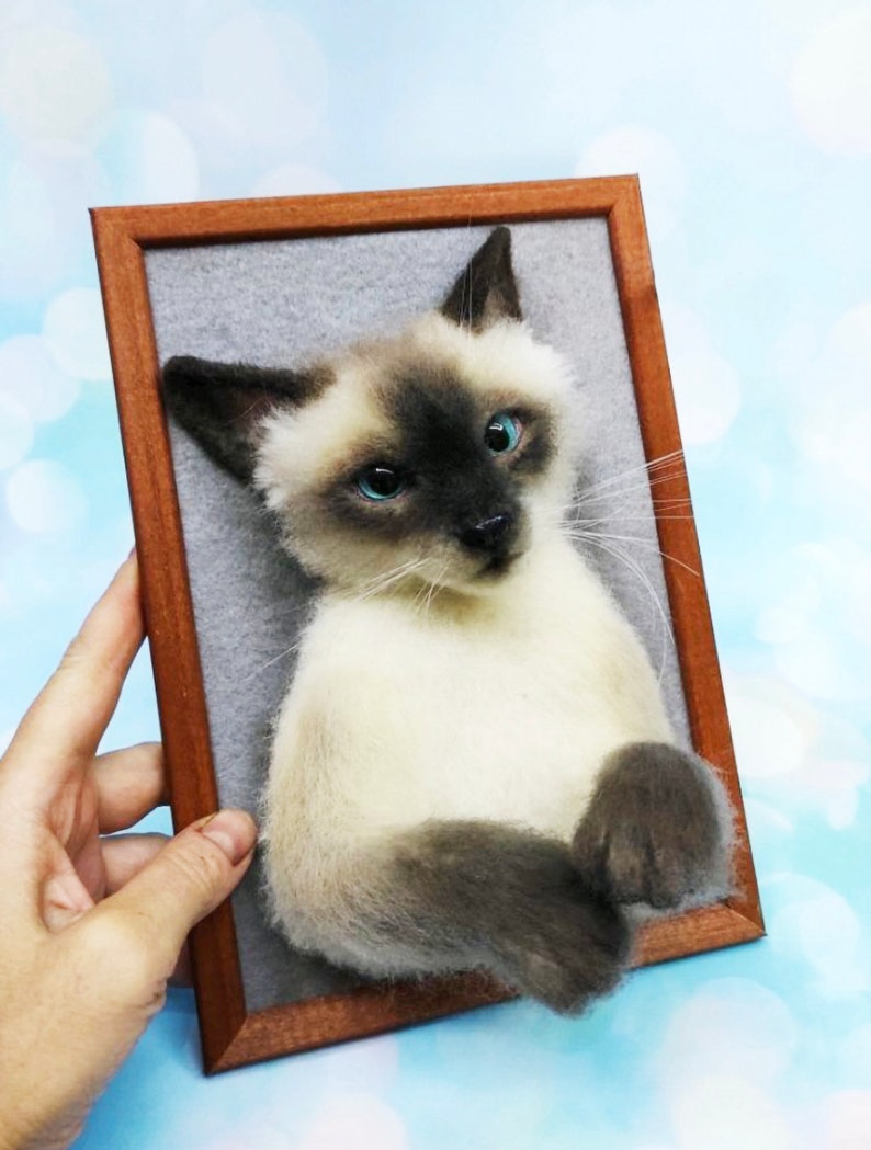 K&ouml;nnte beinhalten: Ein gerahmtes Portr&auml;t einer Siamkatze. Die Katze hat cremefarbenes Fell mit dunkelbraunen Punkten und blauen Augen. Der Holzrahmen hat eine warme braune Farbe. Der Hintergrund ist grau.