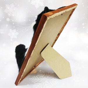 May include: A framed artwork featuring a black and white cat, viewed from the side. The frame is a warm brown, and the artwork is propped up on a cardboard stand. The background has a winter theme with snowflakes.