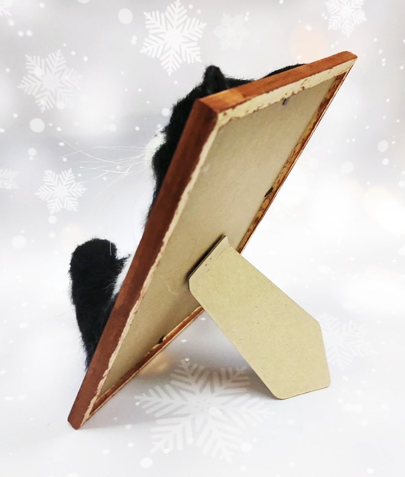 May include: A framed artwork featuring a black and white cat, viewed from the side. The frame is a warm brown, and the artwork is propped up on a cardboard stand. The background has a winter theme with snowflakes.