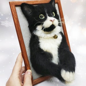 May include: A handmade, framed cat portrait. The cat is black and white with green eyes, a pink nose, and a small bell on its collar. The frame is made of wood. The cat's fur appears soft and textured.
