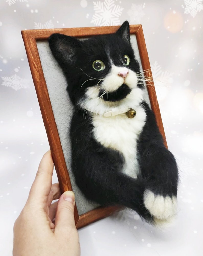 May include: A handmade, framed cat portrait. The cat is black and white with green eyes, a pink nose, and a small bell on its collar. The frame is made of wood. The cat's fur appears soft and textured.