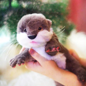 Op de afbeelding: Een klein, pluche otter-speelgoed met bruine vacht, witte borstvacht en roze geverfde klauwen. De otter wordt vastgehouden door een hand.