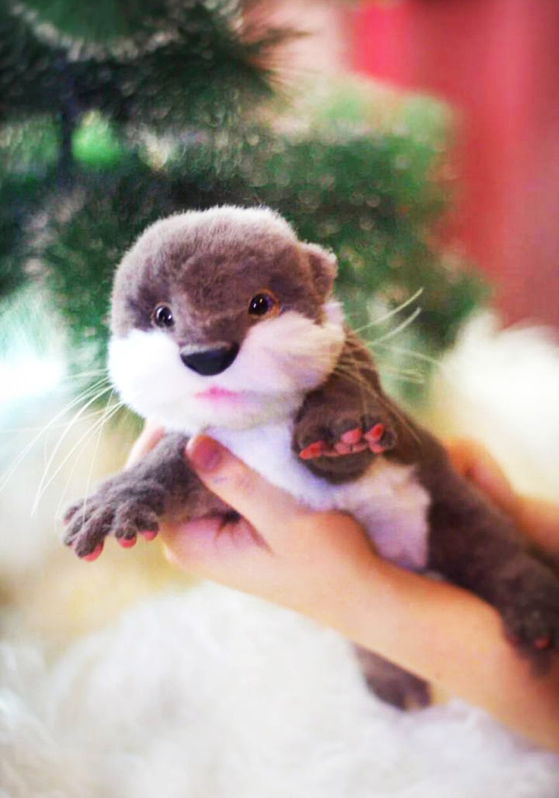 Op de afbeelding: Een klein, pluche otter-speelgoed met bruine vacht, witte borstvacht en roze geverfde klauwen. De otter wordt vastgehouden door een hand.