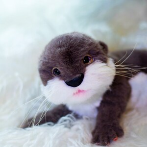 Op de afbeelding: Een pluche grijze en witte otter met zwarte ogen en een roze neus. Het speelgoed ligt op een wit, pluche oppervlak.