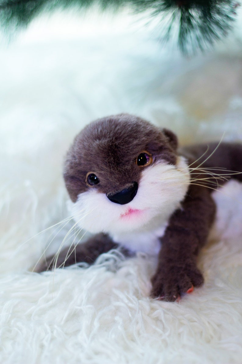 Op de afbeelding: Een pluche grijze en witte otter met zwarte ogen en een roze neus. Het speelgoed ligt op een wit, pluche oppervlak.